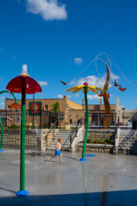 Brightly colored splash pad sprinklers and spouts with a red-headed boy in blue swim trunks running through them. Art sculpture tower with butterflies in the background.