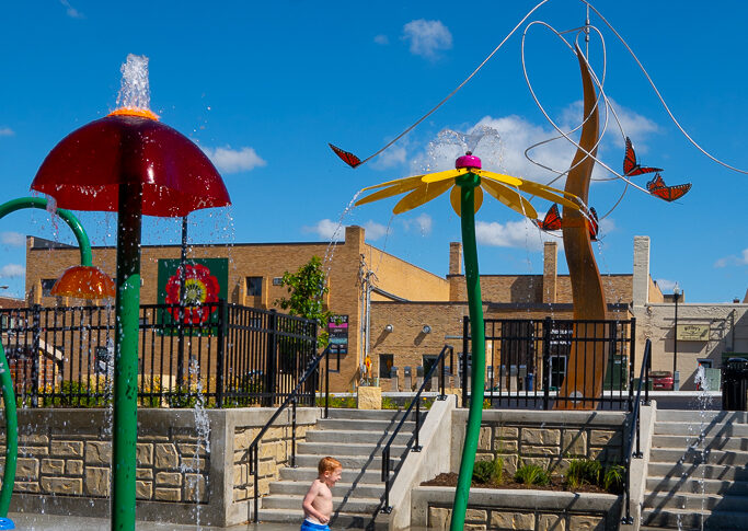 Brightly colored splash pad sprinklers and spouts with a red-headed boy in blue swim trunks running through them. Art sculpture tower with butterflies in the background.