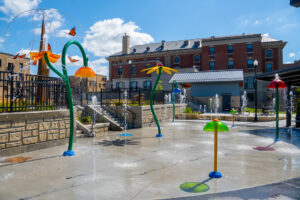 Brightly colored splash pad sprayers and spouts with brick building in background followed by blue sky with clouds.