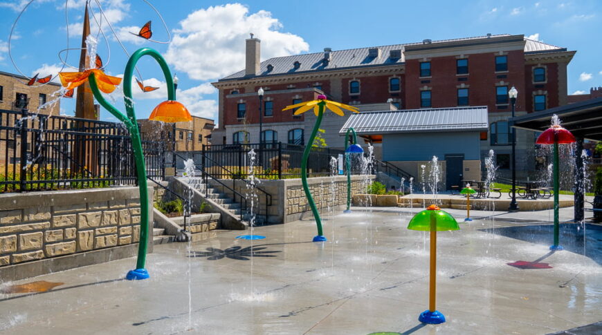 Brightly colored splash pad sprayers and spouts with brick building in background followed by blue sky with clouds.