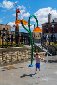 Red-headed young boy in blue swim trunks runs through splash pad sprinkler with butterfly sculpture tower in the background.