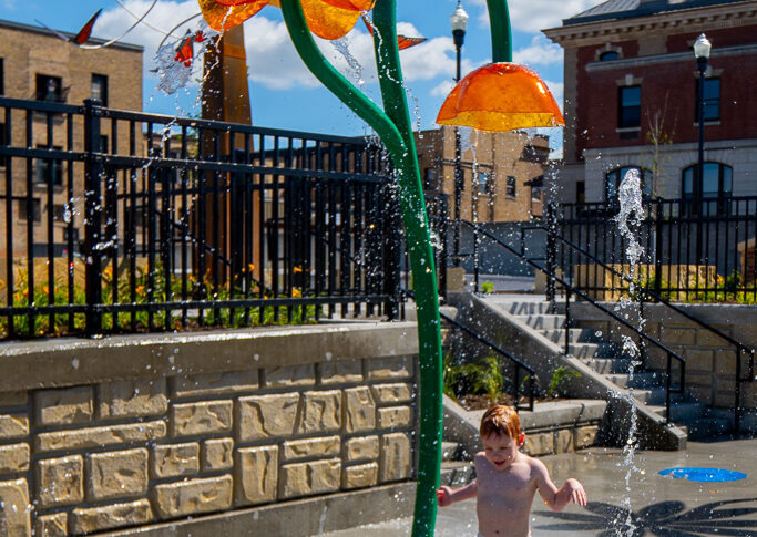 Red-headed young boy in blue swim trunks runs through splash pad sprinkler with butterfly sculpture tower in the background.
