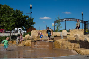 Waterfall rock feature of Riverfront splash pad with toddlers playing and adult in background watching.