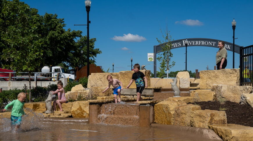 Waterfall rock feature of Riverfront splash pad with toddlers playing and adult in background watching.