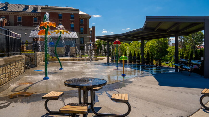Riverfront Splashpad flower water features with covered shelter and benches to the right and picnic tables to the foreground.