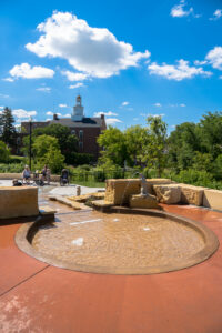 Circular wading pool with small waterfall down to splash pad with green trees in background and city hall building behind.