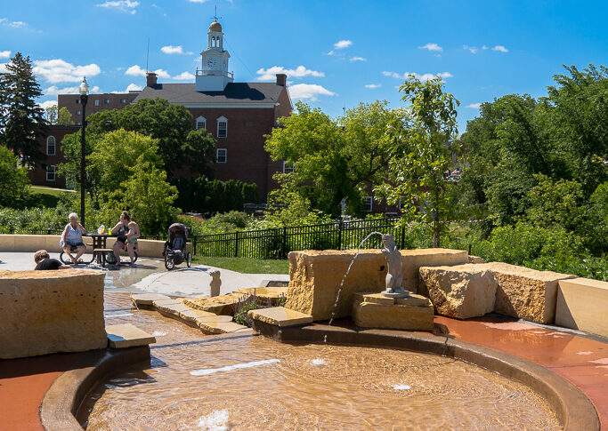 Circular wading pool with small waterfall down to splash pad with green trees in background and city hall building behind.