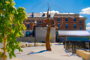 Tall rust-colored sculpture with orange butterflies hanging down it with brick industrial building in background.