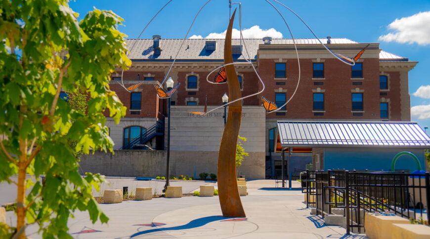 Tall rust-colored sculpture with orange butterflies hanging down it with brick industrial building in background.