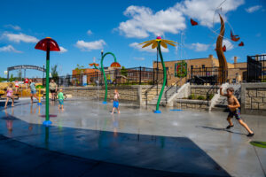 Splash pad sprinklers and sprayers in flower and mushroom themed with kids running around playing underneath with bright blue sky and white clouds behind and downtown Fergus Falls buildings.