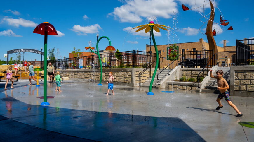 Splash pad sprinklers and sprayers in flower and mushroom themed with kids running around playing underneath with bright blue sky and white clouds behind and downtown Fergus Falls buildings.
