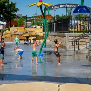 Brightly colored splash pad sprayers and spouts with children in swimsuits running underneath with the "Spies Riverfront Park" sign in the background.