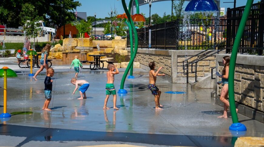 Brightly colored splash pad sprayers and spouts with children in swimsuits running underneath with the "Spies Riverfront Park" sign in the background.