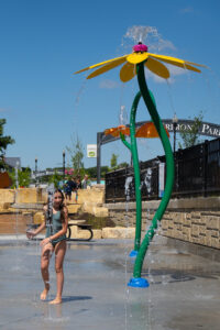 Young girl with long brown hair in olive green one-piece swimsuit standing underneath a yellow flower splash pad sprayer.