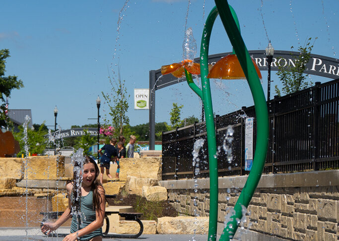 Young girl with long brown hair in olive green one-piece swimsuit standing underneath a yellow flower splash pad sprayer.