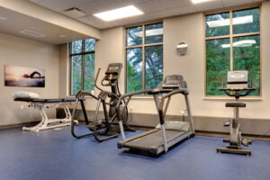 Rehabilitation exercise machines and adjustment table with three tall windows overlooking lush green trees behind.