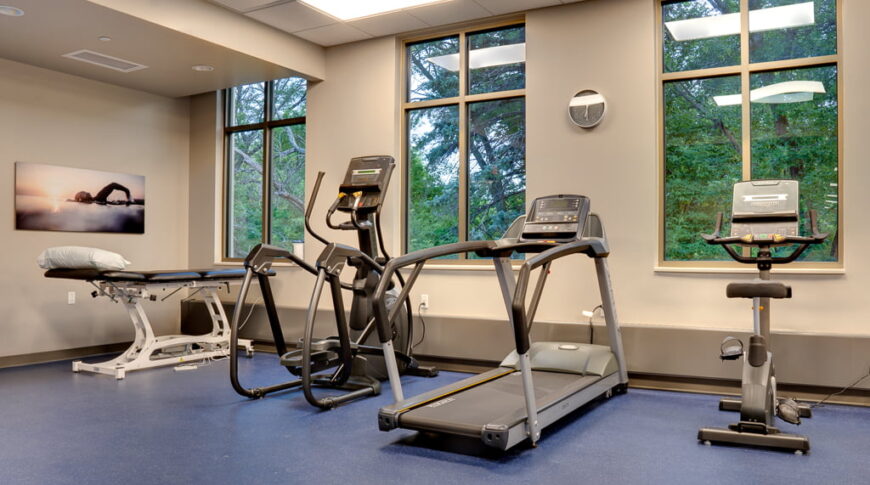 Rehabilitation exercise machines and adjustment table with three tall windows overlooking lush green trees behind.