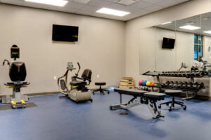 Rehab physical therapy gym with two machines on the back beige wall with free weights stacked on a stand to the right wall. Blue durable flooring underneath.