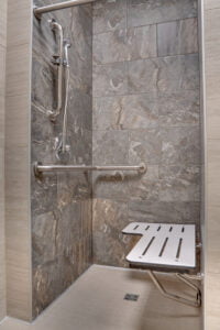 Accessible shower stall with fold-down bench, hand raiils, and hand-held showerhead attachment. Large format gray and beige tiles surround.