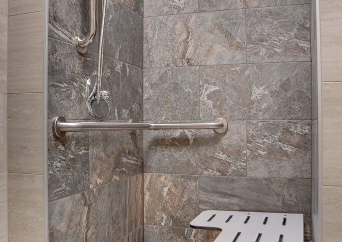 Accessible shower stall with fold-down bench, hand raiils, and hand-held showerhead attachment. Large format gray and beige tiles surround.