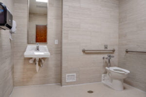 Large, accessible bathroom with single white hanging pedestal sink to the back left, mirror atop with soap dispenser and towel dispenser to the left. Toilet with accessibility bars to the right. Entire room flanked with large-format gray tiles with smaller square tiles on the floor.