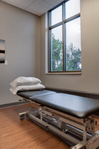 Physical therapy office exam room with adjustment table in the middle with a couple pillows topped on top. Brown laminate faux-wood flooring underneath, greige-painted walls with large double window overlooking green trees.
