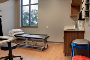 Physical therapy exam room fitted with adjustment table, workstation with desktop computer monitor and rolling swivel stool. Cabinetry with supplies and garbages for medical waste to the far right wall. Double window overlooking green trees to the back wall.