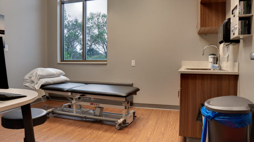Physical therapy exam room fitted with adjustment table, workstation with desktop computer monitor and rolling swivel stool. Cabinetry with supplies and garbages for medical waste to the far right wall. Double window overlooking green trees to the back wall.