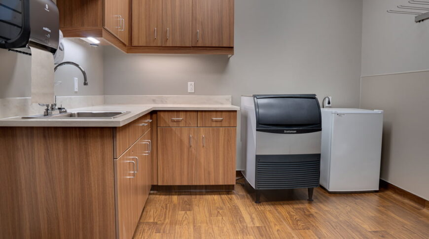 Physical therapy backroom with light faux-wood cabinetry in L-shape to the back and left walls with sink and paper towel dispenser. Ice machine and mini fridge to the back right.