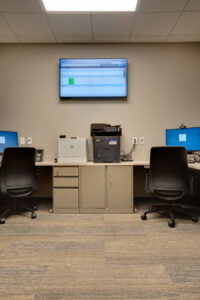 Physical therapy collaborative office with two workstations in the back corner, printing station in the middle with cabinet files underneath and tv monitor mounted on the wall behind.