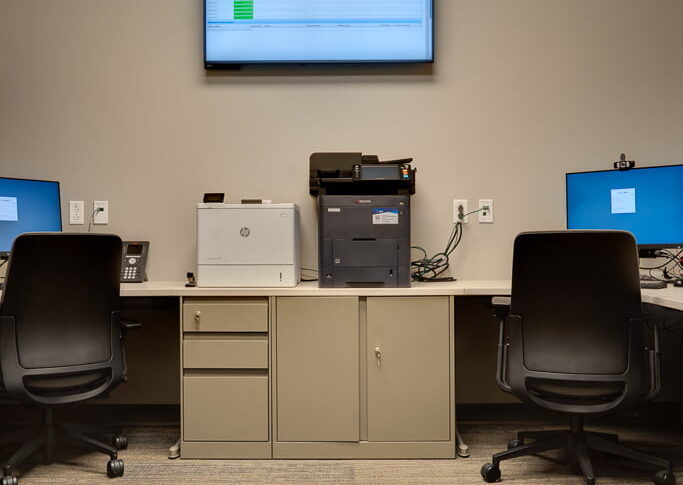 Physical therapy collaborative office with two workstations in the back corner, printing station in the middle with cabinet files underneath and tv monitor mounted on the wall behind.