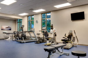 Physical therapy rehabilitation training room with exercise machines lining the back wall overlooking the green trees through three tall windows. Adjustment table on the back wall.
