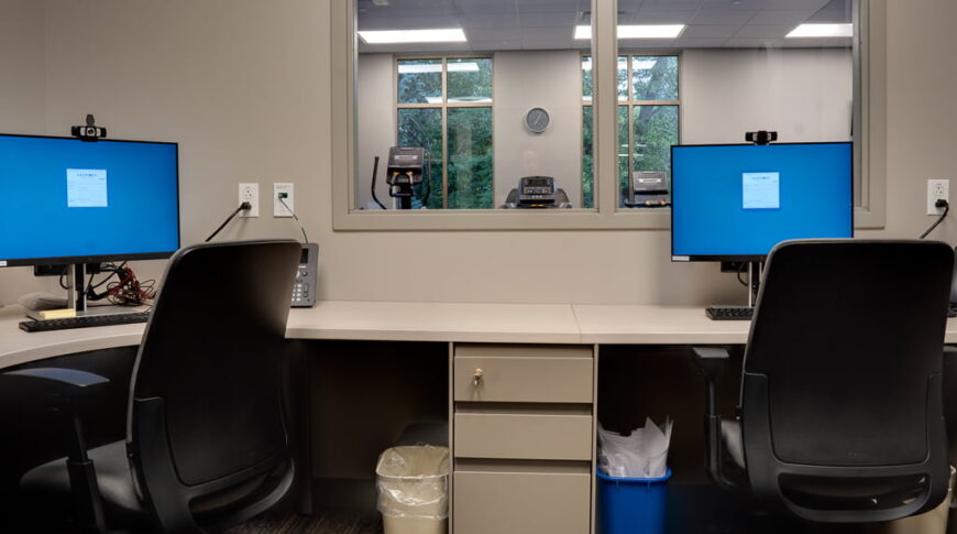 Physical therapy rehab office with see-through window overlooking the therapy gym. Two workstations sharing a back counter with file cabinetry in between.