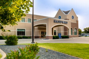 Exterior of beige-colored stucco building with porte-cochère with the word "Sanford" in white letters atop. Blue sky in background and green grass in front with shrubs, trees, and grass for landscaping.