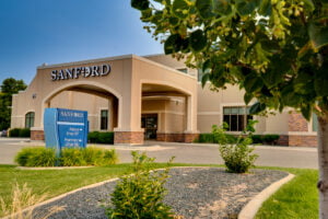 Exterior of beige-colored stucco building with porte-cochère with the word "Sanford" in white letters atop. Blue sky in background and green grass in front with shrubs, trees, and grass for landscaping.