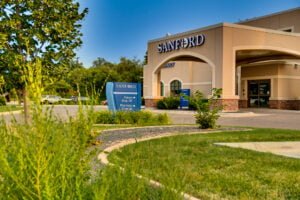 Exterior of beige-colored stucco building with porte-cochère with the word "Sanford" in white letters atop. Blue sky in background and green grass in front with shrubs, trees, and grass for landscaping at Sanford West Fargo Clinic.