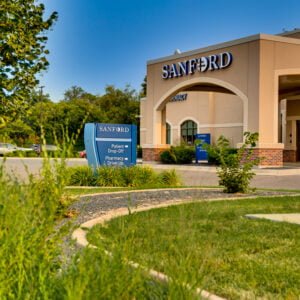Exterior of beige-colored stucco building with porte-cochère with the word "Sanford" in white letters atop. Blue sky in background and green grass in front with shrubs, trees, and grass for landscaping at Sanford West Fargo Clinic.