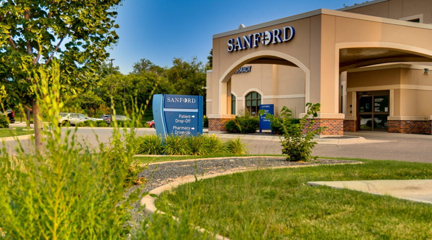Exterior of beige-colored stucco building with porte-cochère with the word "Sanford" in white letters atop. Blue sky in background and green grass in front with shrubs, trees, and grass for landscaping at Sanford West Fargo Clinic.