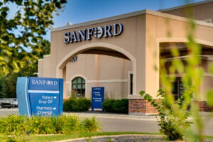 Exterior of beige-colored stucco building with porte-cochère with the word "Sanford" in white letters atop. Blue sky in background and green grass in front with shrubs, trees, and grass for landscaping at Sanford West Fargo clinic.
