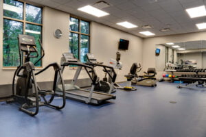 Beige-colored exercise therapy room with two tall windows overlooking greenery outside with exercise machines besides. Back wall of mirrors with free weight rack to the far back right wall.