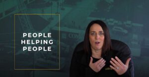Woman with medium-colored skin, blue eyes and dark brown shoulder-length hair speaking with hands talking while sitting in office chair. Behind is a darn green picture overlayed background of a construction site. To the left side is a yellow outlined box with the words "People Helping People" inside.