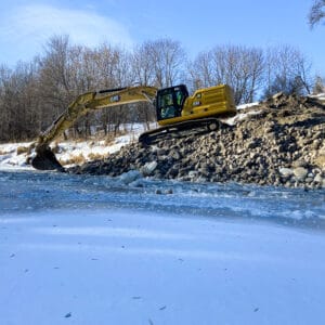Excavator moving rocks alongside an ice-coated riverbank to cover stream barbs and prevent erosion.