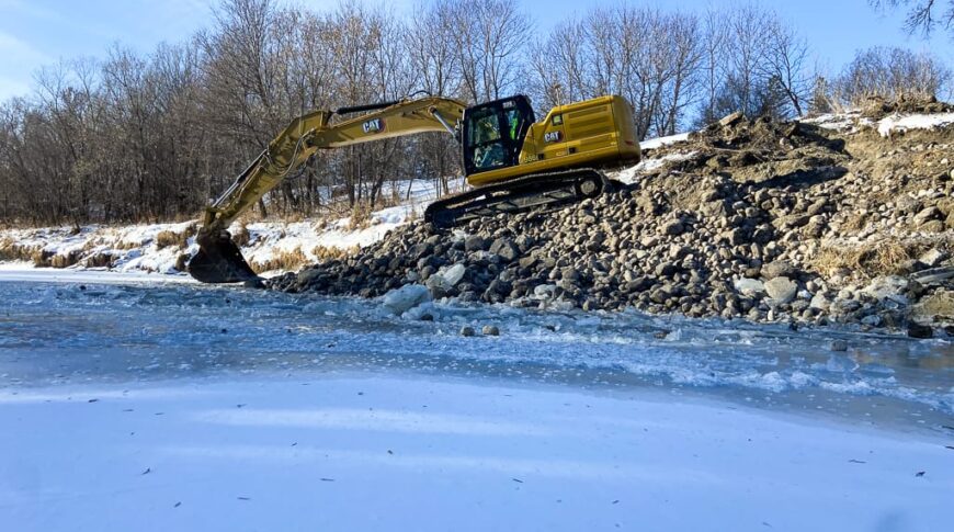 Excavator moving rocks alongside an ice-coated riverbank to cover stream barbs and prevent erosion.