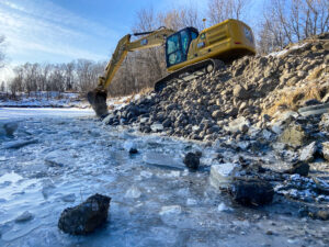 Excavator moving rocks alongside an ice-coated riverbank to cover stream barbs and prevent erosion.