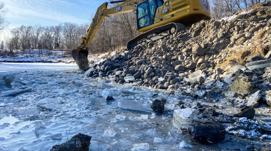 Excavator moving rocks alongside an ice-coated riverbank to cover stream barbs and prevent erosion.