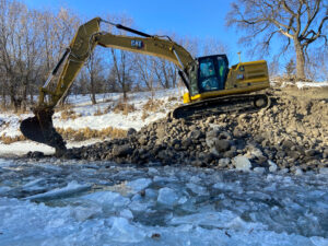 Excavator moving rocks alongside an ice-coated riverbank.