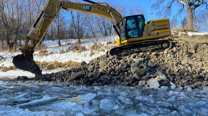 Excavator moving rocks alongside an ice-coated riverbank.
