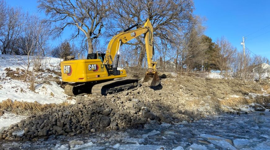 Yellow excavator moving rocks along a riverbank during wintertime.