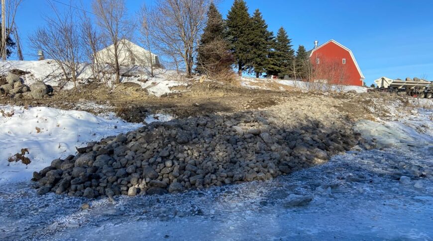 Rip Rap Rocks covering Stream Barbs on a riverbank with red barn and white building in background.