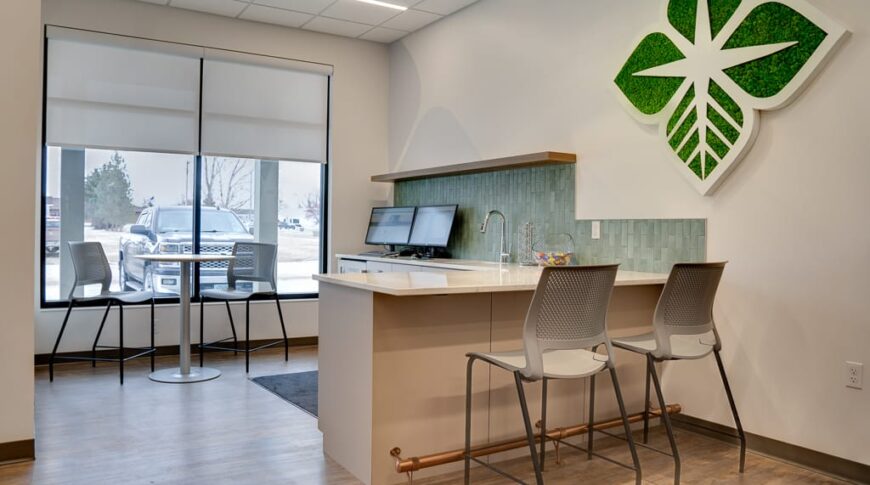 A minibar for refreshments and a customer-use computer at the front entry of AgCountry Lamoure, ND. Branding throughout with moss-covered logo on the wall, green accent pendant lights, and green tile backsplash.